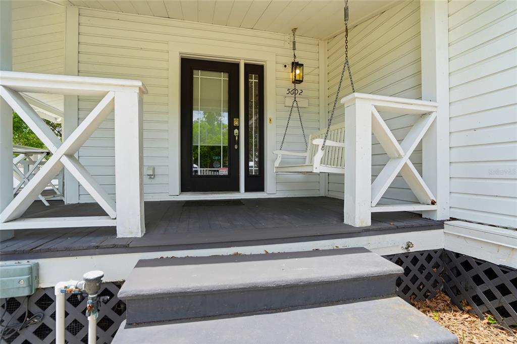A front porch with a white swing, wooden railing, and a dark-framed glass door — 5602 N OLA AVENUE, TAMPA, FL