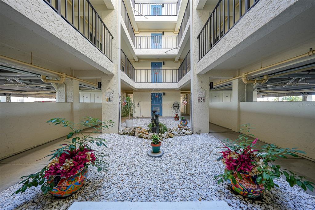 An outdoor courtyard features a gravel ground, a small fountain, and potted plants between buildings — 1701 BEACH ROAD UNIT 202, ENGLEWOOD, FL