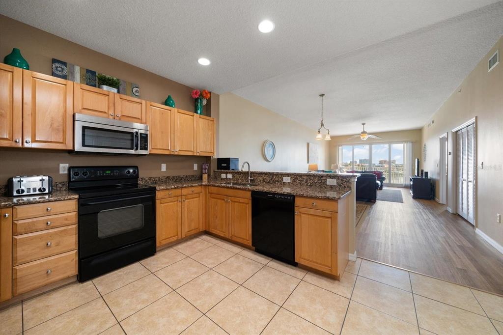 A kitchen with light wood cabinets, granite countertops, and black appliances opens to a living area — 530 S GULFVIEW BOULEVARD UNIT 401, CLEARWATER BEACH, FL