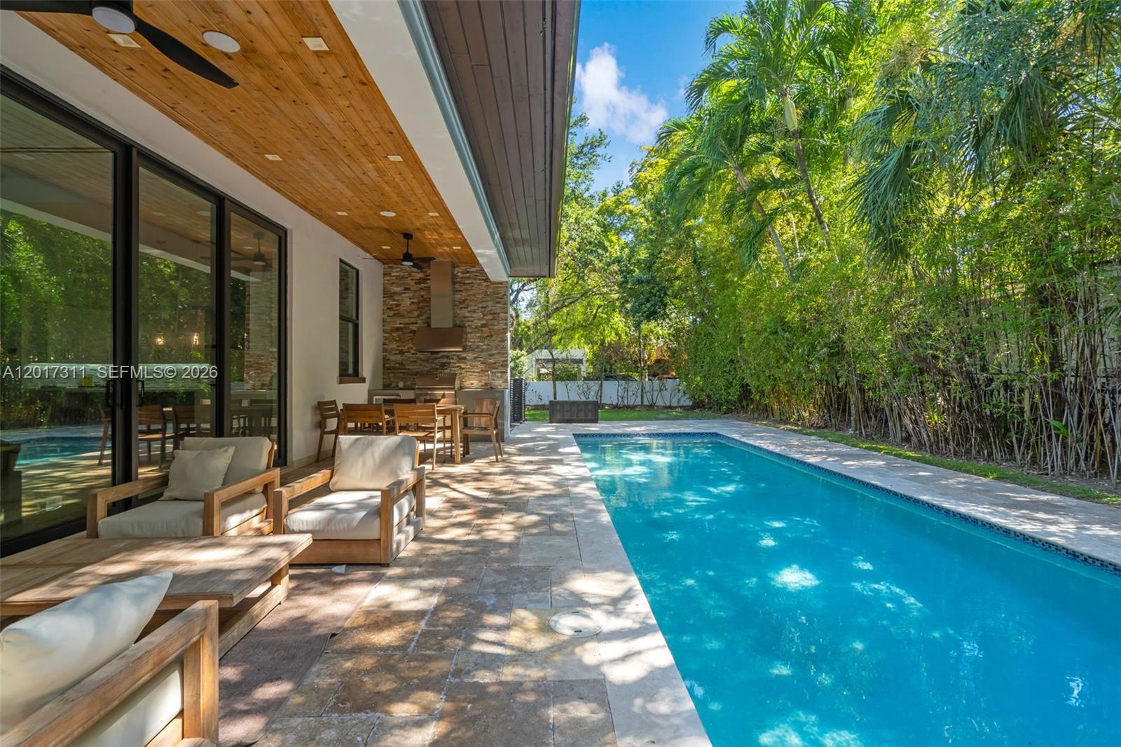 A covered patio with a stone outdoor kitchen overlooks a rectangular swimming pool and garden — 1010 Genoa St, Coral Gables FL 33134, CORAL GABLES, FL