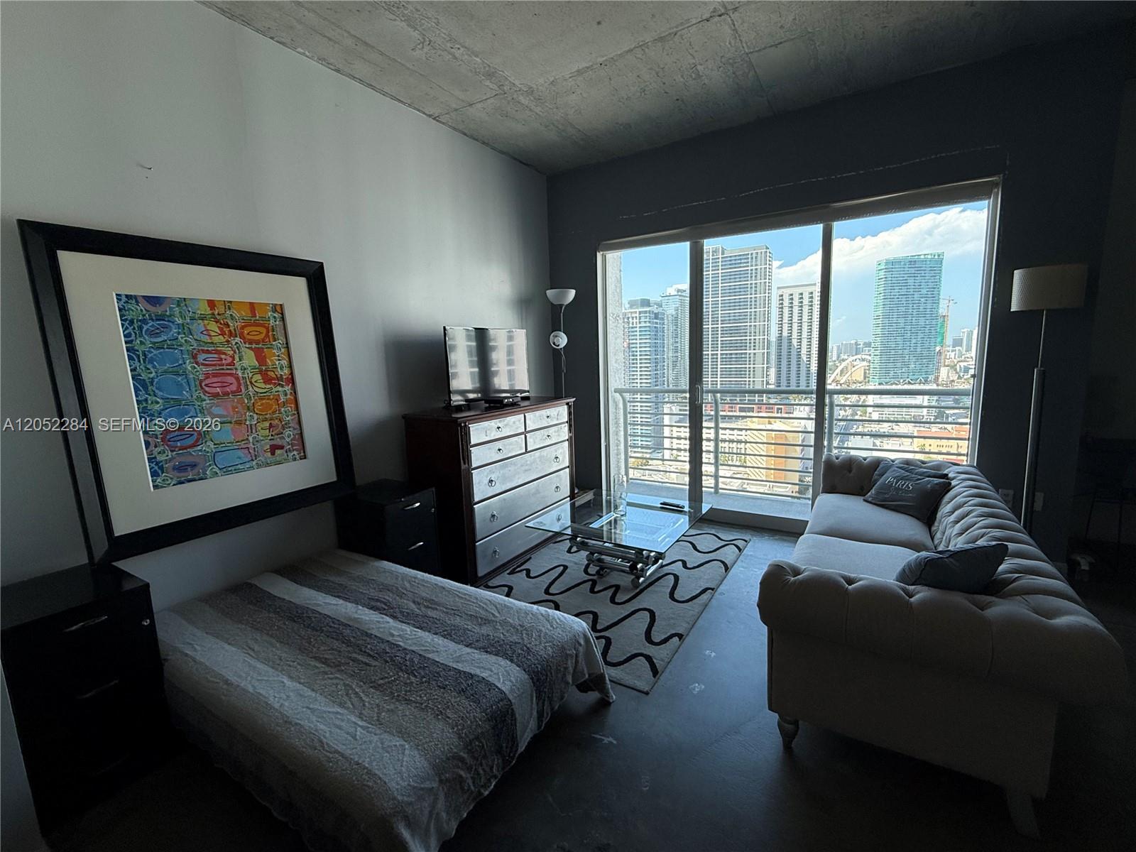 A studio apartment features a bed, sofa, dresser, and floor-to-ceiling windows overlooking a city skyline — 133 NE 2nd Ave # 2811, Miami FL 33132, MIAMI, FL