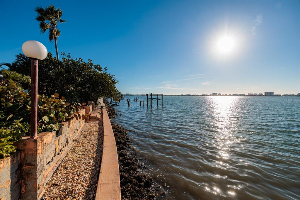A stone walkway runs alongside a seawall overlooking a sunlit bay with distant buildings — 1844 VENETIAN POINT DRIVE, CLEARWATER, FL