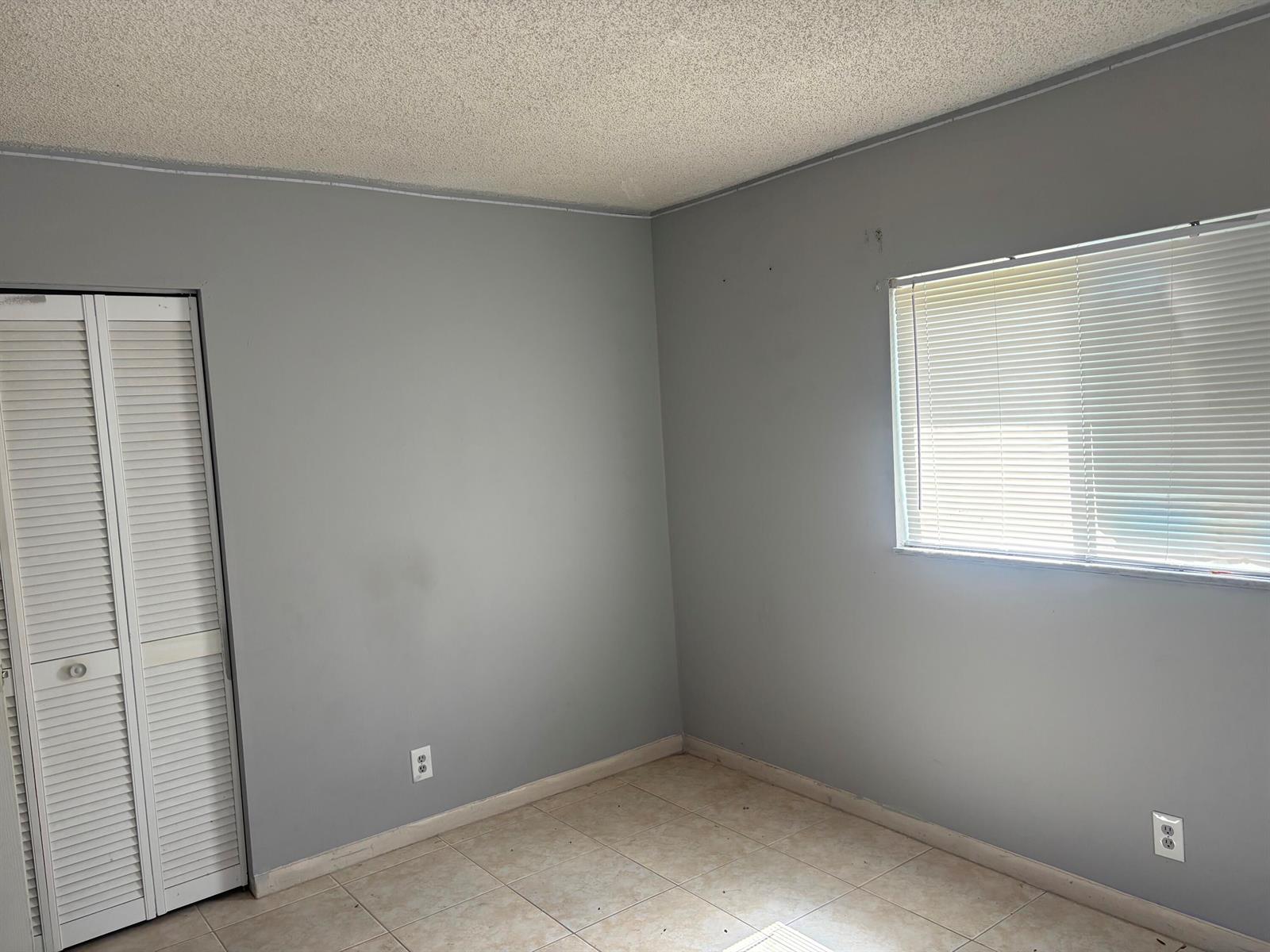An empty bedroom with gray walls, tile flooring, a white louvered closet door, and a window — 1250 SW 5TH Terrace, Deerfield Beach FL 33441, DEERFIELD BEACH, FL