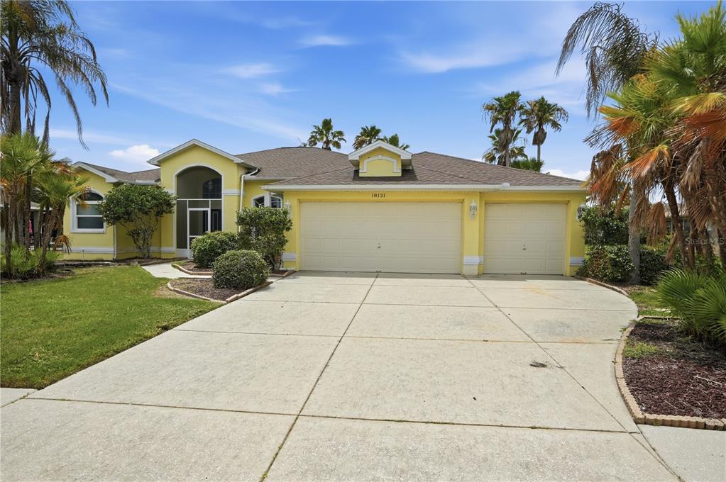 A yellow single-story house features a three-car garage, a concrete driveway, and palm trees — 18131 RIVARD BOULEVARD, BROOKSVILLE, FL