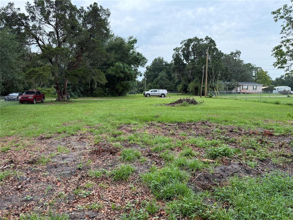 A grassy, partially cleared lot features scattered debris, mature trees, and parked vehicles in the background — 5427 SE 137TH LANE, SUMMERFIELD, FL