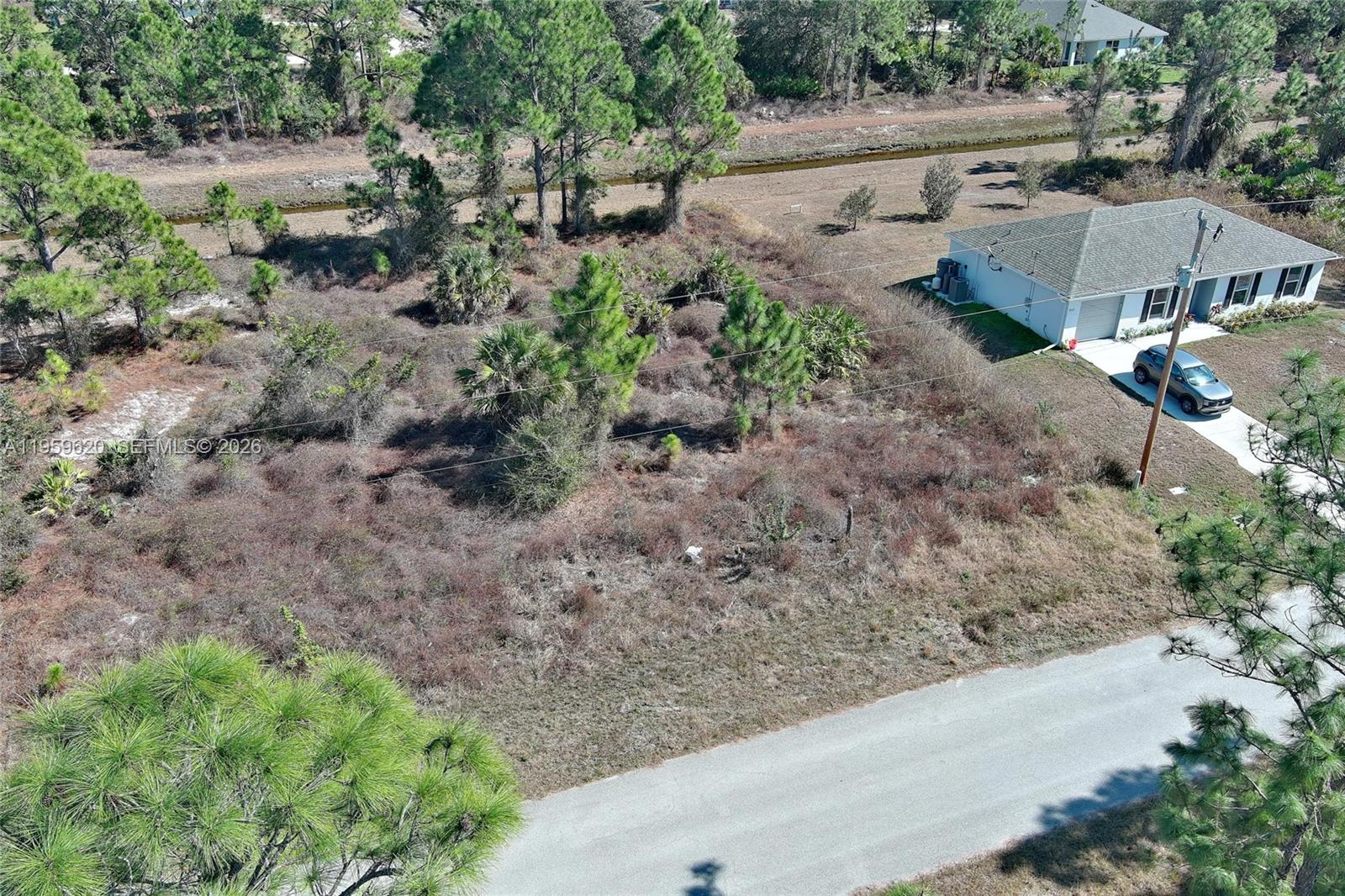 An aerial view shows a vacant, brush-covered lot adjacent to a single-story house and road — 943 FOXDALE AVE, Lehigh Acres FL 33974, LEHIGH ACRES, FL