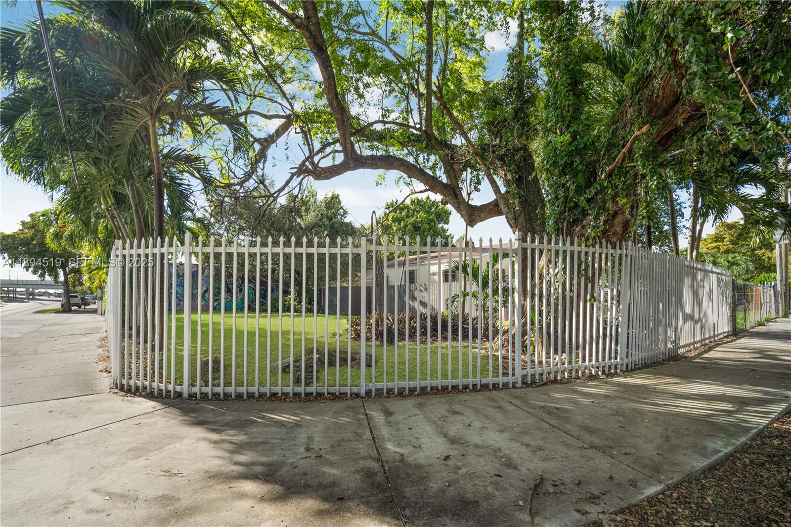 A white metal fence encloses a grassy yard with large trees and a house behind — 1121 NW 29th St, Miami FL 33127, MIAMI, FL