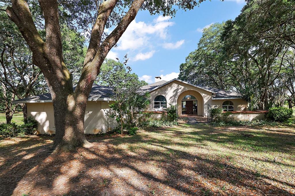 A single-story house with a stucco exterior and arched entryway sits among large oak trees — 27011 SE HIGHWAY 42, UMATILLA, FL