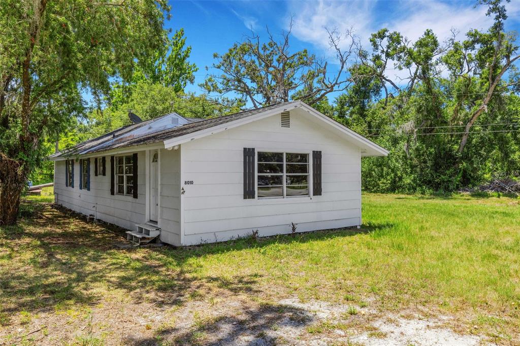 A single-story white house with black shutters sits on a grassy lot with trees — 8010 BROWER DRIVE, BRADENTON, FL