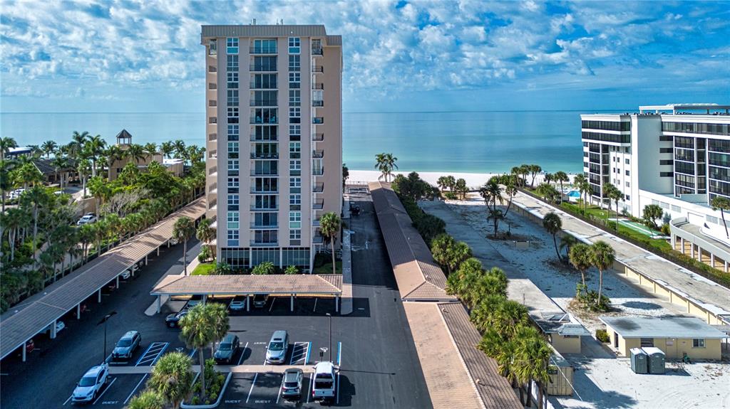 An aerial view shows a tall residential building, a parking lot, and a sandy beach — 1212 BENJAMIN FRANKLIN DRIVE UNIT 607, SARASOTA, FL