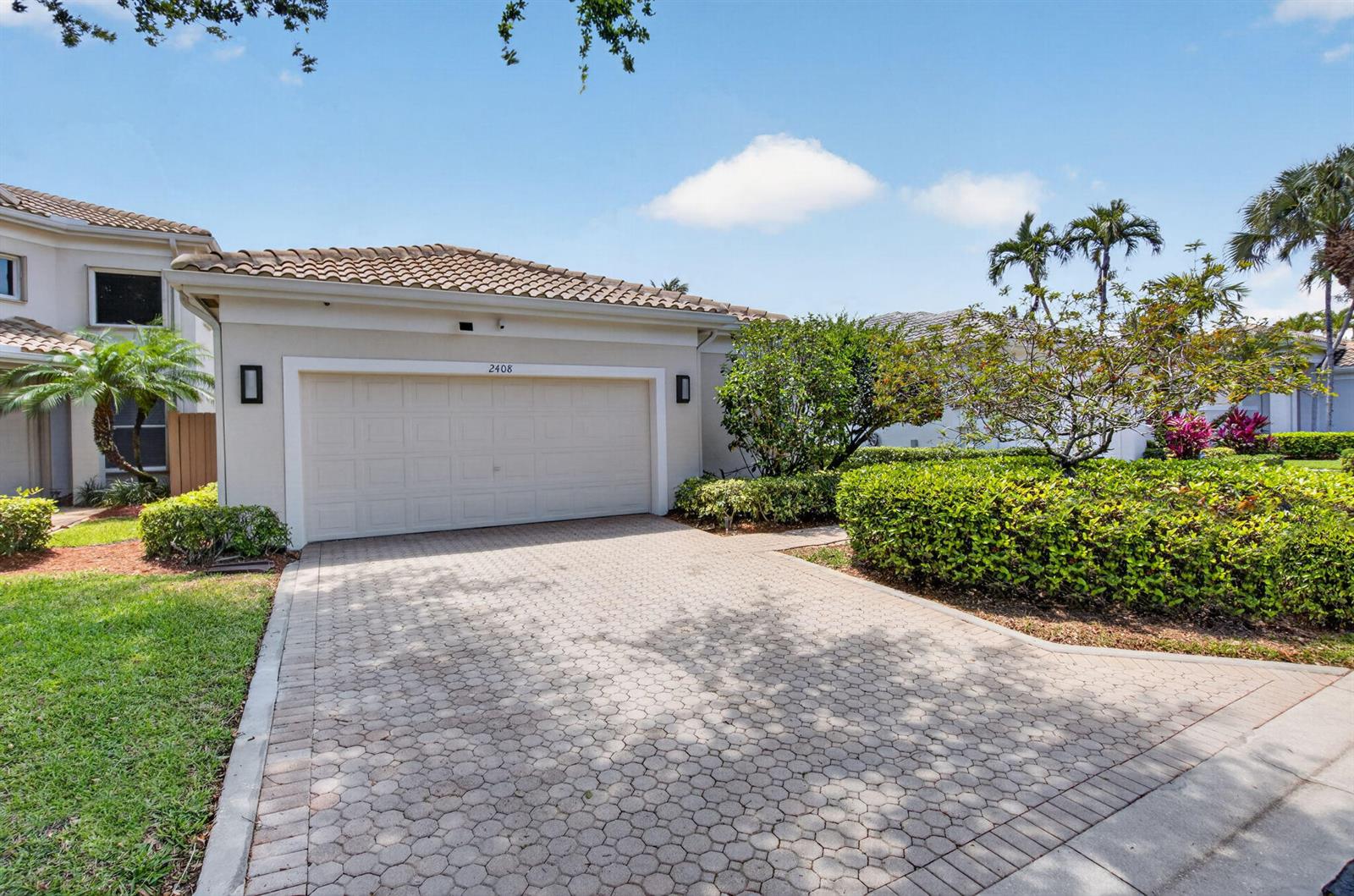 A beige house exterior features a two-car garage, a paved driveway, and manicured landscaping — 2408 NW 67th Street, Boca Raton FL 33496, BOCA RATON, FL