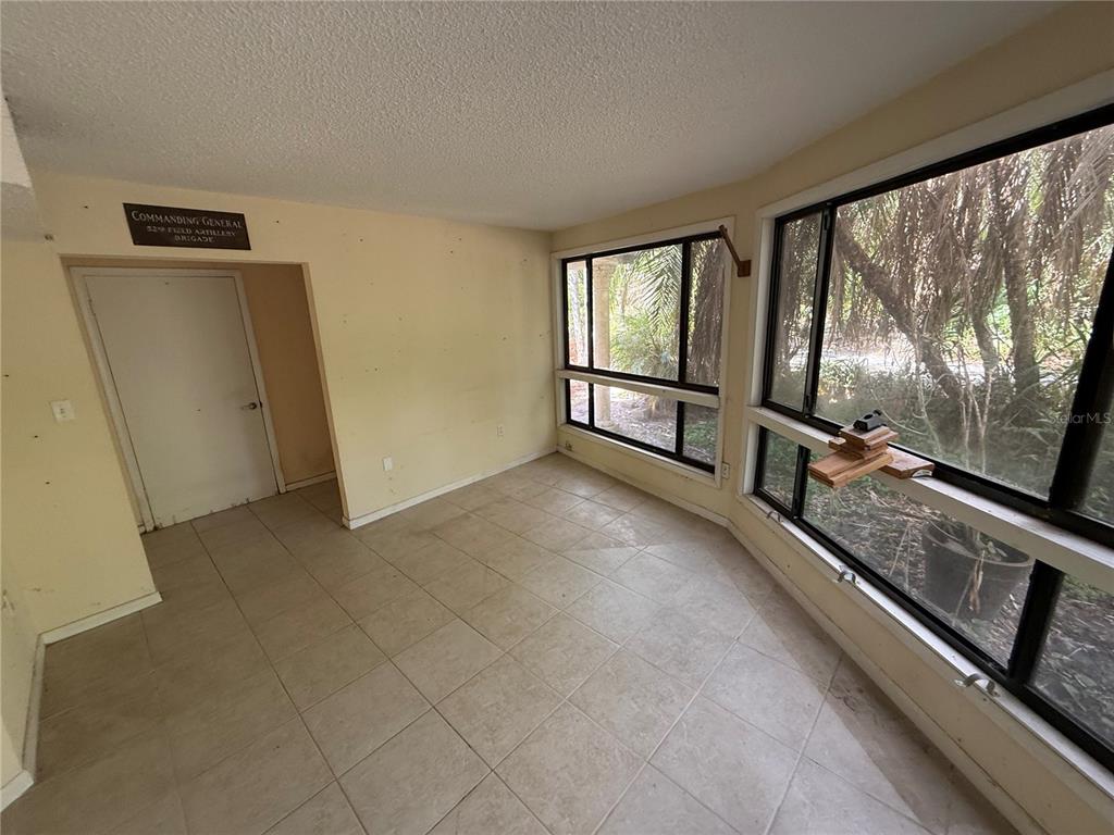 An empty room with light-colored tile flooring, beige walls, and large windows overlooking trees — 6904 PINETREE LANE E, LAKELAND, FL