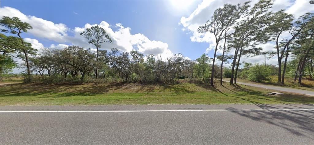 A paved road runs in front of a vacant, tree-covered lot under a bright sky — COMMONWEALTH AVENUE N, POLK CITY, FL