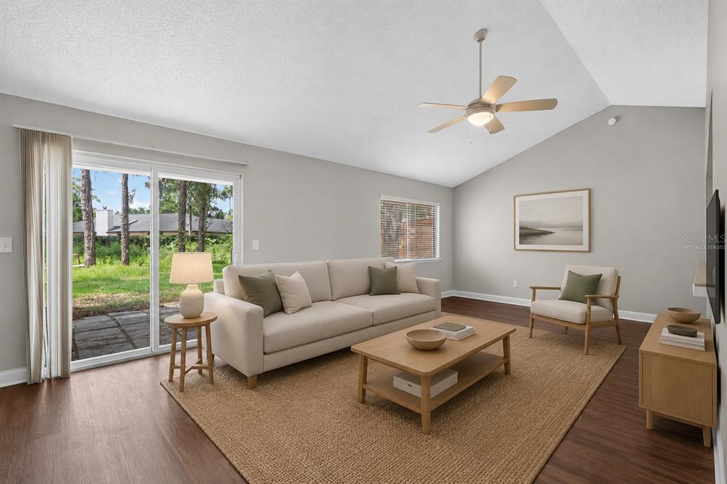 This virtually staged living room features a vaulted ceiling, wood-look flooring, and a sliding glass door — 3918 PINE BREEZE ROAD S, JACKSONVILLE, FL