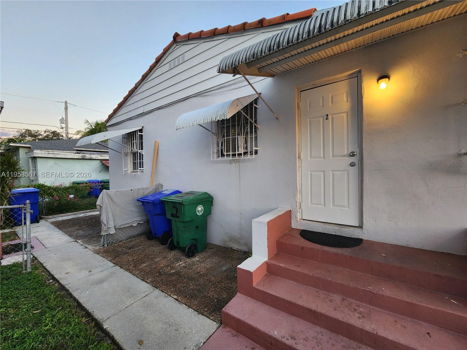 The exterior of a light-colored house features a white door, red steps, and trash bins — 405 NW 64th St # 2, Miami FL 33150, MIAMI, FL