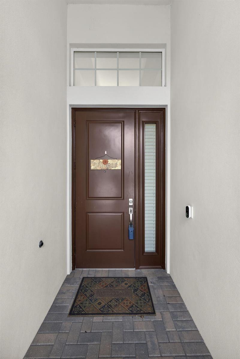 A brown front door with a side light and transom window sits on a paved porch — 1411 Thistle Place, Loxahatchee FL 33470, LOXAHATCHEE, FL