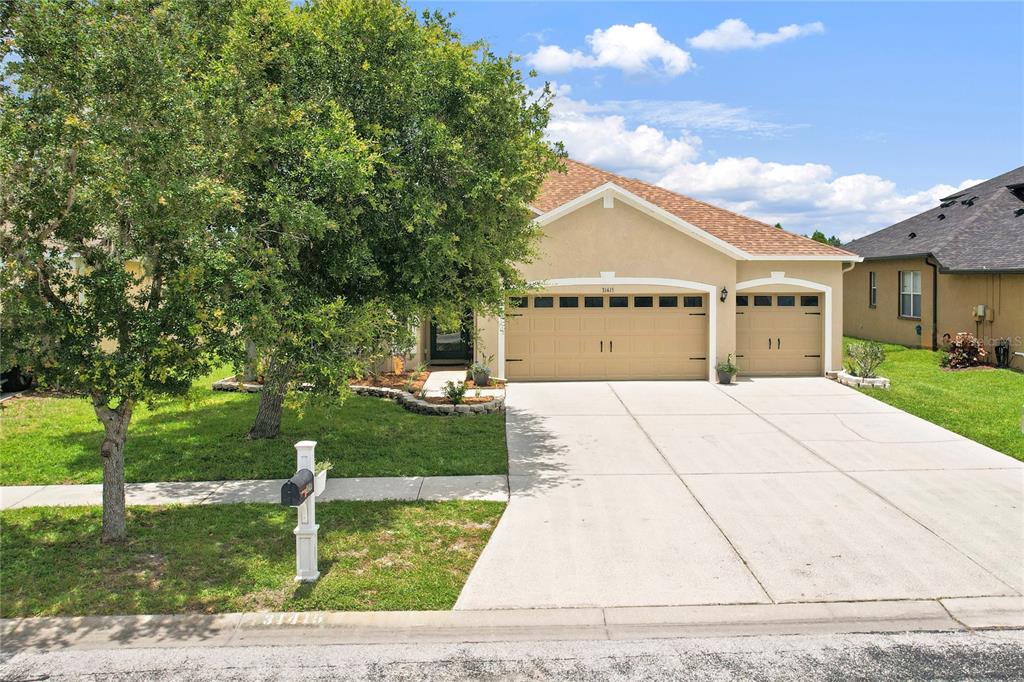 A tan, single-story suburban house features a three-car garage and a large front yard tree — 31415 BRIDGEGATE DRIVE, WESLEY CHAPEL, FL