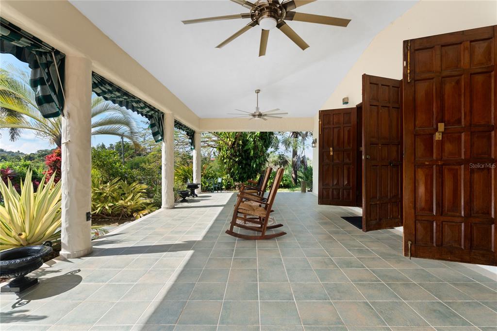 A covered patio features tiled flooring, rocking chairs, ceiling fans, and large wooden doors — 59 REINA ISABEL, RIO GRANDE, PR