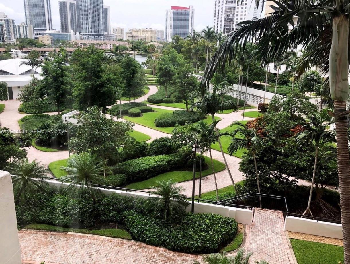 An elevated view of a landscaped courtyard with paved walkways, lush greenery, and distant skyscrapers — 600 Three Islands Blvd # 418, Hallandale Beach FL 33009, HALLANDALE BEACH, FL