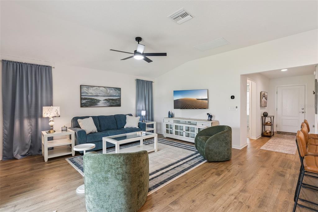 A living room features a blue sofa, two green armchairs, and a wall-mounted television — 7608 DANIEL HEARNS WAY, THE VILLAGES, FL