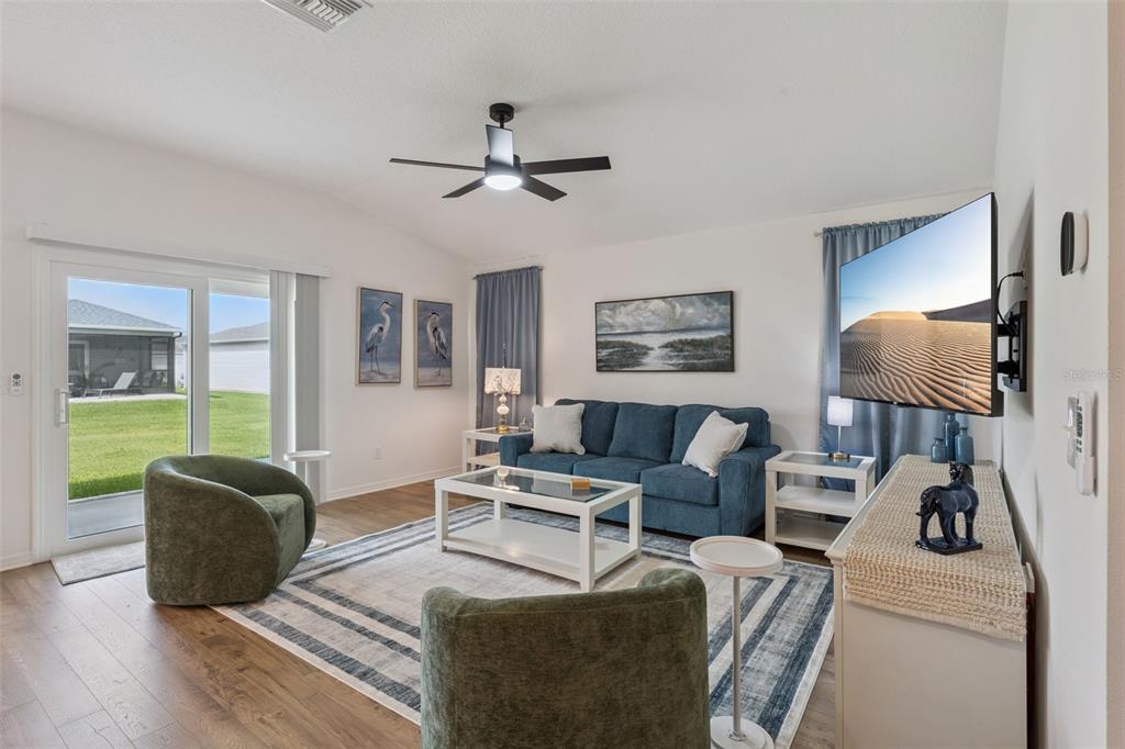A living room features a blue sofa, two green armchairs, a television, and sliding doors — 7608 DANIEL HEARNS WAY, THE VILLAGES, FL