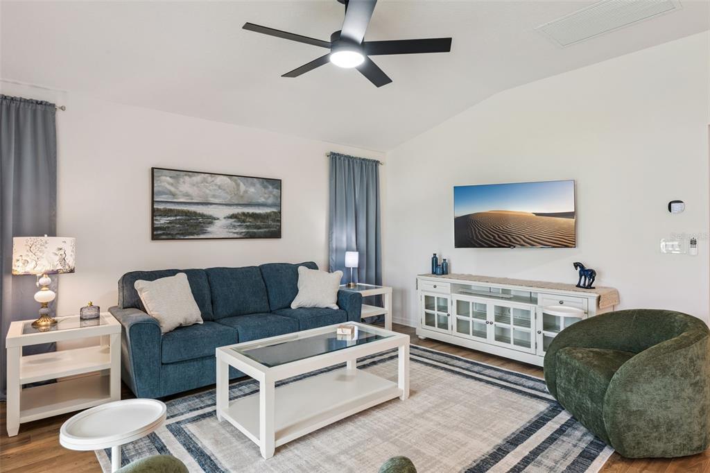 A living room features a blue sofa, white coffee table, media console, and ceiling fan — 7608 DANIEL HEARNS WAY, THE VILLAGES, FL