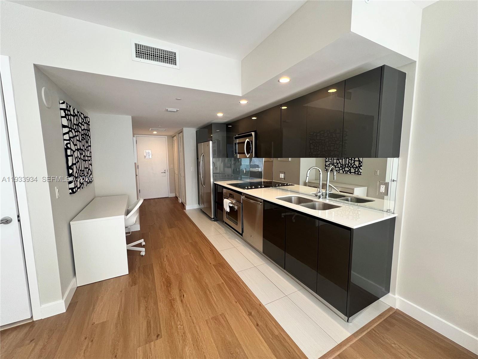 A modern kitchen features dark glossy cabinets, stainless steel appliances, and a white desk area — 31 SE 6th St # 703, Miami FL 33131, MIAMI, FL
