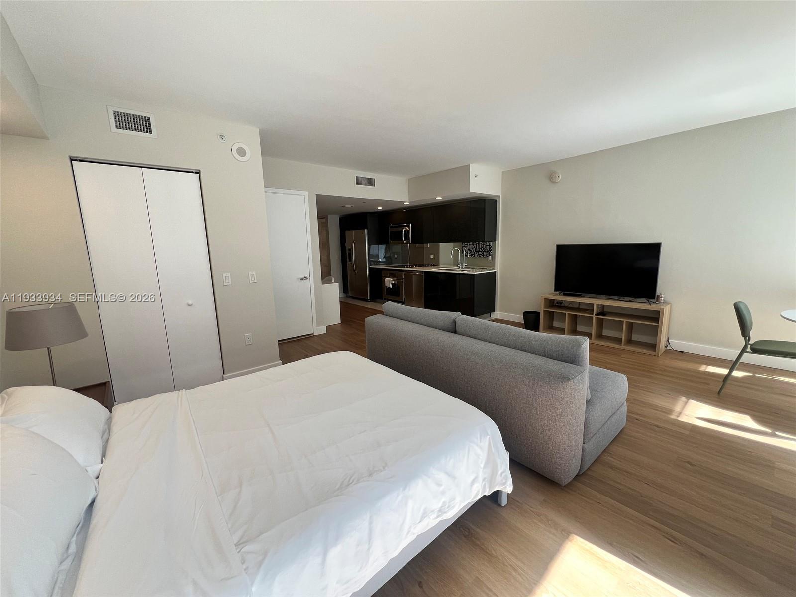 A studio apartment features a bed, a gray sofa, a television, and a kitchen area — 31 SE 6th St # 703, Miami FL 33131, MIAMI, FL