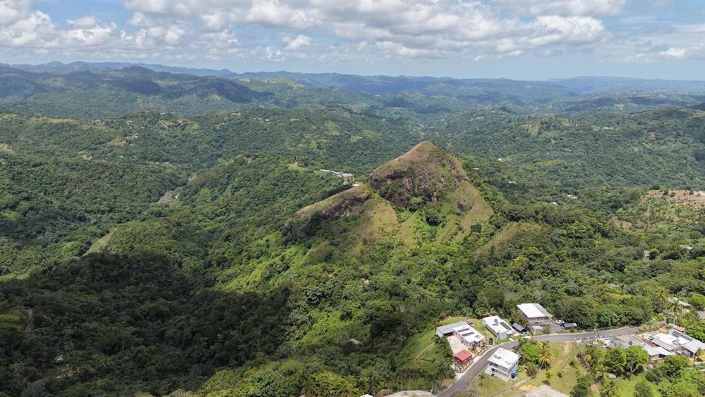 An aerial view shows a prominent rocky hill surrounded by lush green mountains and houses — CAMINO VITORIANO, LARES, PR