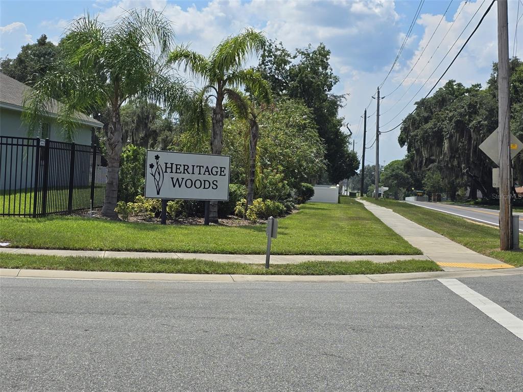A sign for Heritage Woods stands on a grassy corner next to a sidewalk — 605 ARIA DR., LAKELAND, FL