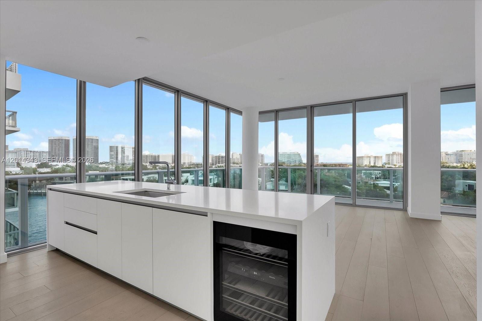 A modern kitchen island with a wine cooler sits before floor-to-ceiling windows overlooking a city — 9201 E Bay Harbor, Bay Harbor Islands FL 33154, BAY HARBOR ISLANDS, FL