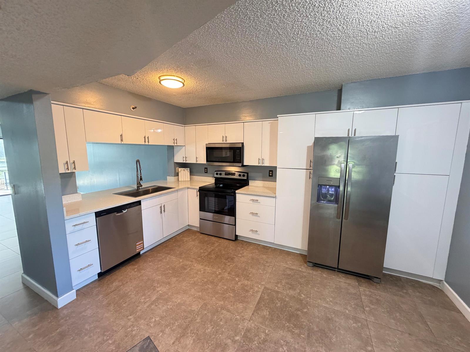 A kitchen features white cabinetry, stainless steel appliances, light countertops, and brown tile flooring — 2202 S Cypress Bend Drive # 101, Pompano Beach FL 33069, POMPANO BEACH, FL