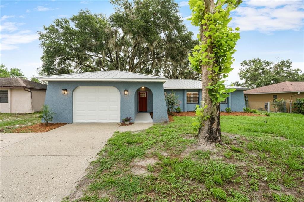 A single-story blue house with a white garage door, metal roof, and a large tree — 12830 LAKE TREE LANE, HUDSON, FL