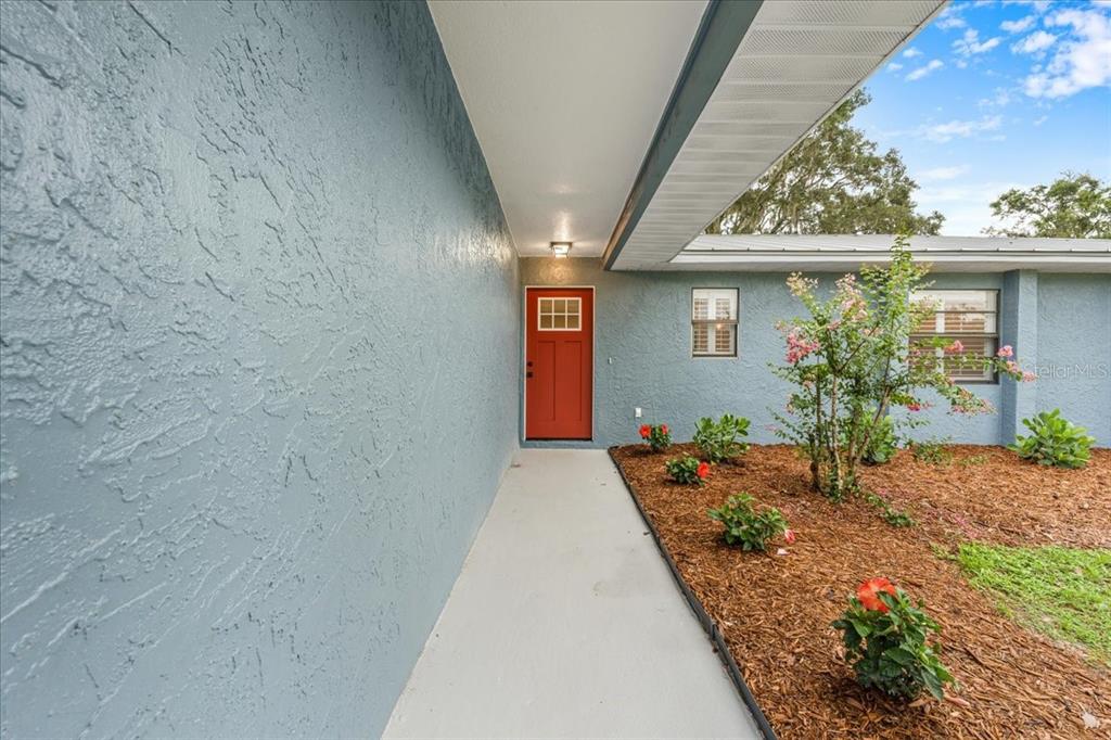A covered concrete walkway leads to a red front door on a blue stucco house — 12830 LAKE TREE LANE, HUDSON, FL