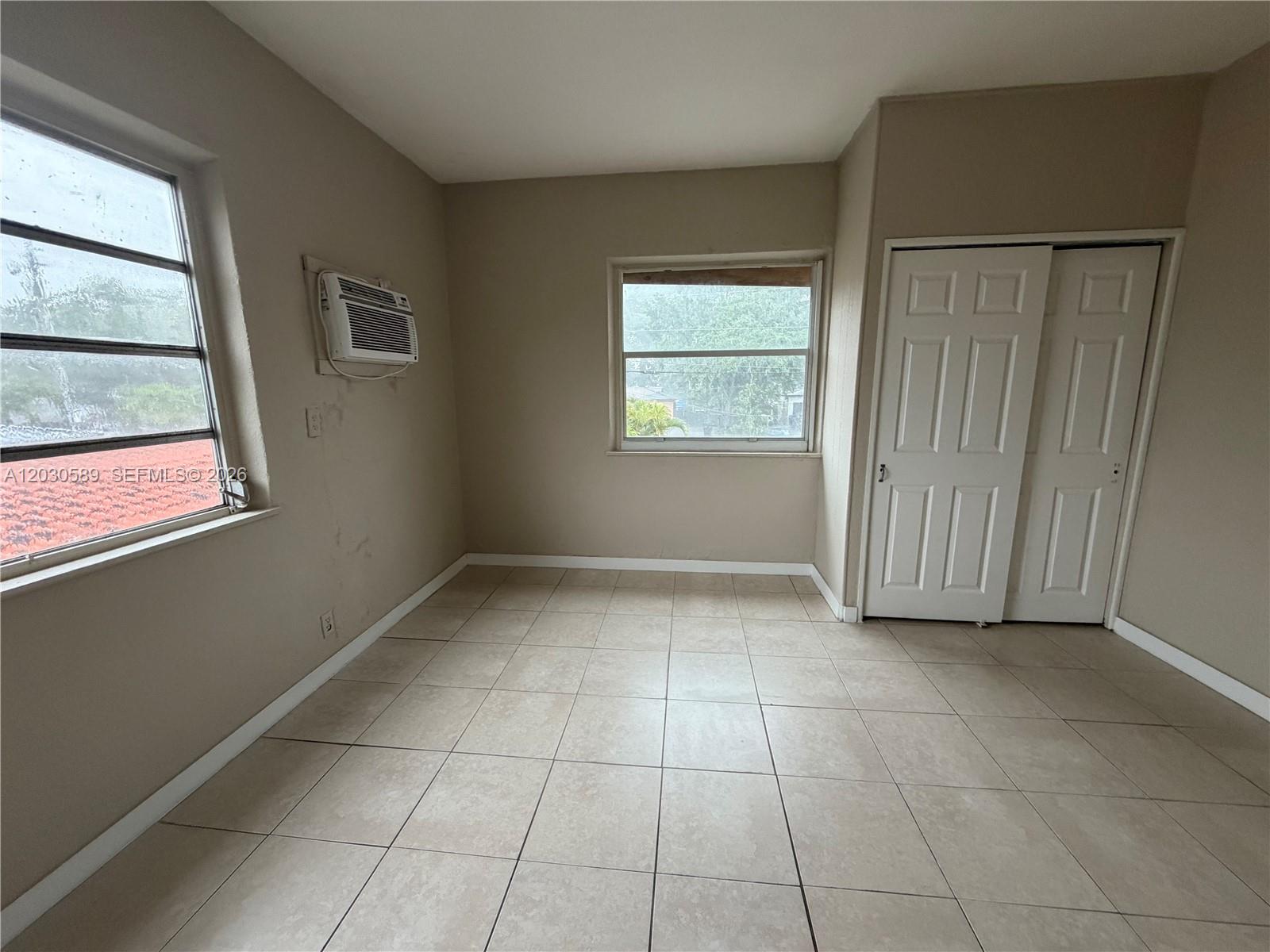 A bedroom with beige walls, tiled flooring, two windows, a wall-mounted air conditioner, and closets — 1422 NW 26th Ave, Miami FL 33125, MIAMI, FL