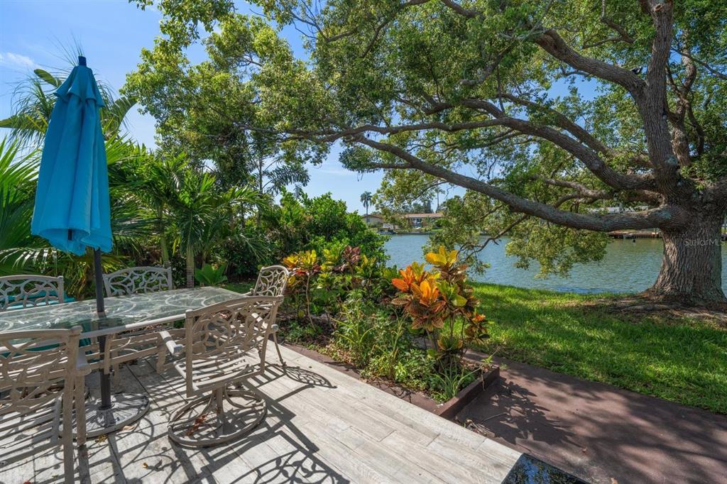 A patio with a dining set and umbrella overlooks a lake under a large tree — 4114 SUNRAY DRIVE, HOLIDAY, FL