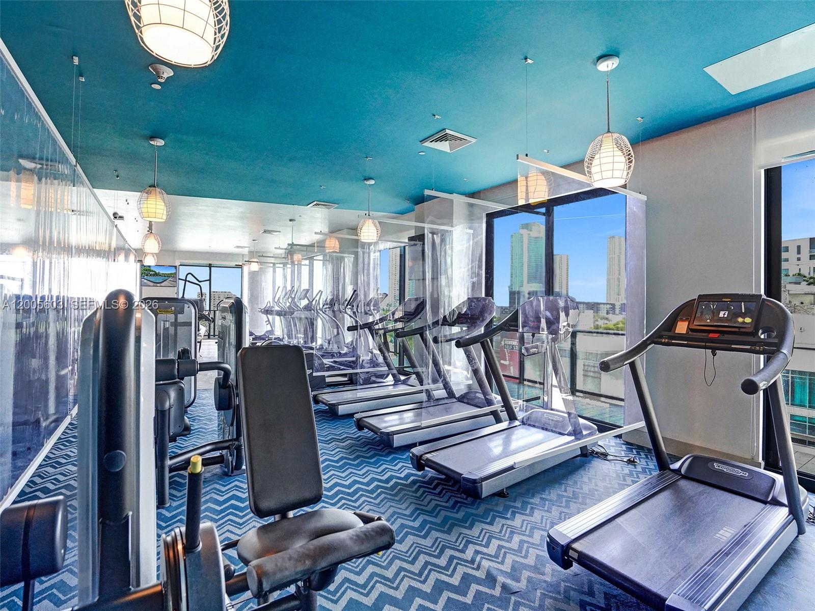 A gym with treadmills, weight equipment, blue chevron carpet, and floor-to-ceiling windows overlooking a city — 121 NE 34th St # 1806A, Miami FL 33137, MIAMI, FL