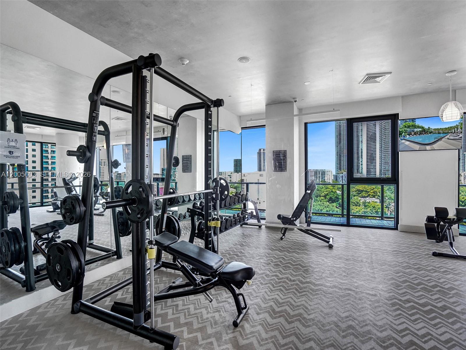 A gym with a weight rack, adjustable benches, and floor-to-ceiling windows overlooking a city skyline — 121 NE 34th St # 1806A, Miami FL 33137, MIAMI, FL