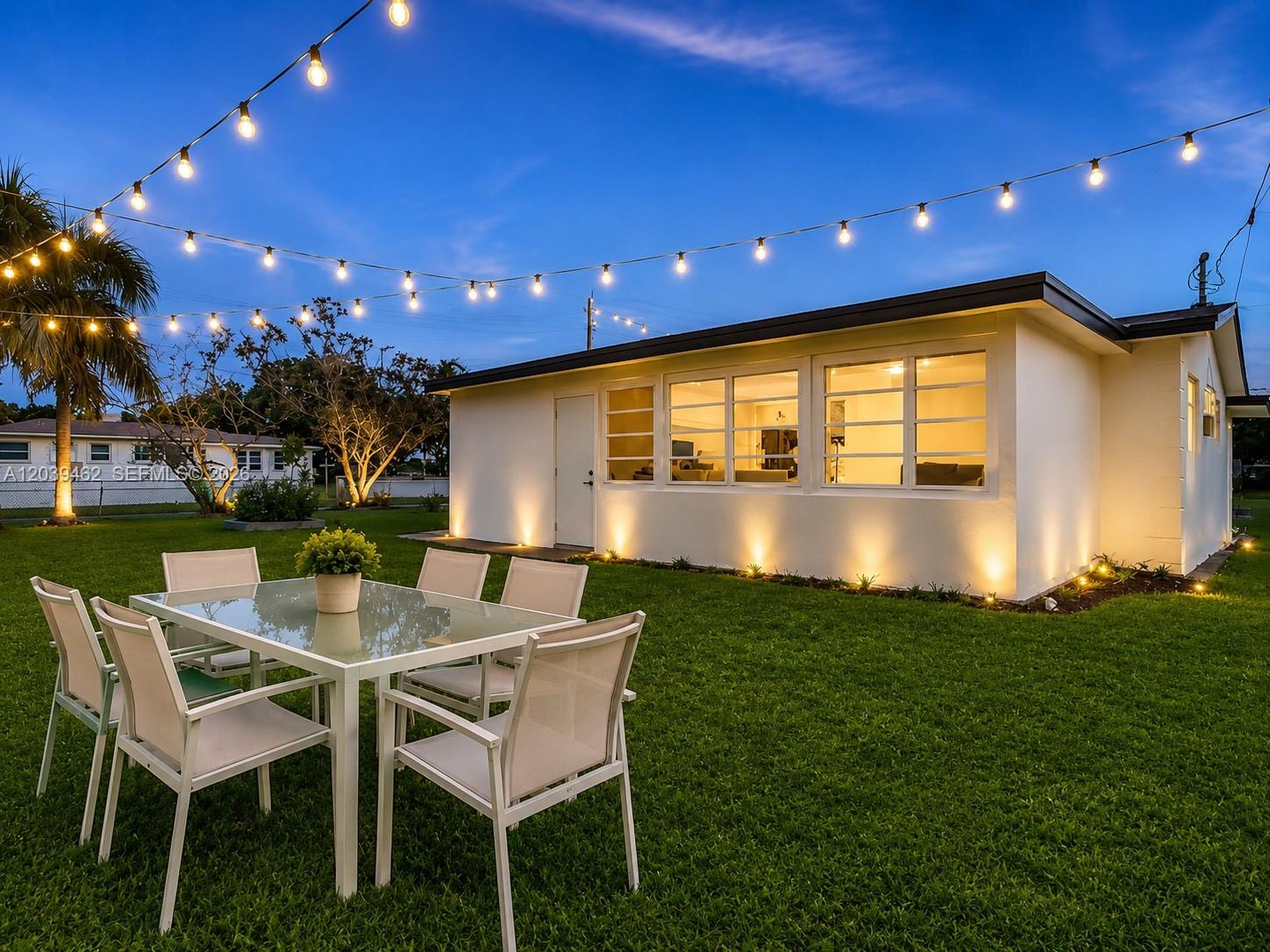 A backyard patio features a glass-topped dining set illuminated by overhead string lights at dusk — 1220 NE 8 St, Pompano Beach FL 33060, POMPANO BEACH, FL