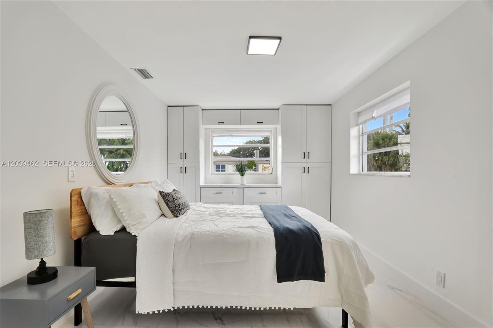 A bedroom features a bed, built-in white cabinetry around a window, and a round mirror — 1220 NE 8 St, Pompano Beach FL 33060, POMPANO BEACH, FL