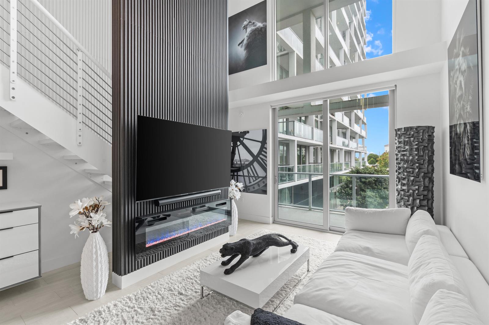 A modern living room features a slatted accent wall with a fireplace, television, and balcony — 2000 Metropica Way # 214, Sunrise FL 33323, SUNRISE, FL
