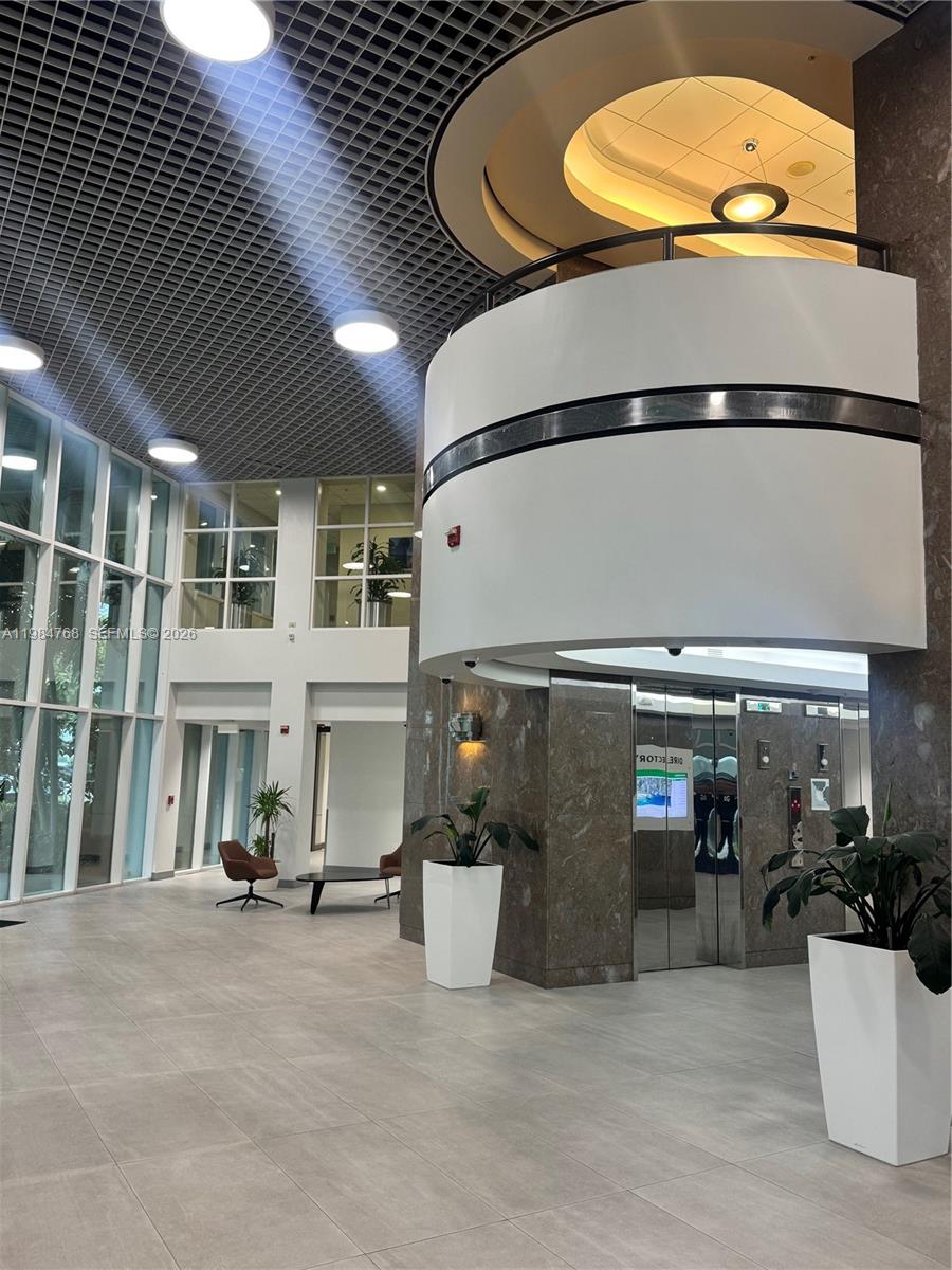 A modern office building lobby features a curved balcony, glass elevator, and tiled flooring — 5959 Waterford District Dr # 101, Miami FL 33126, MIAMI, FL