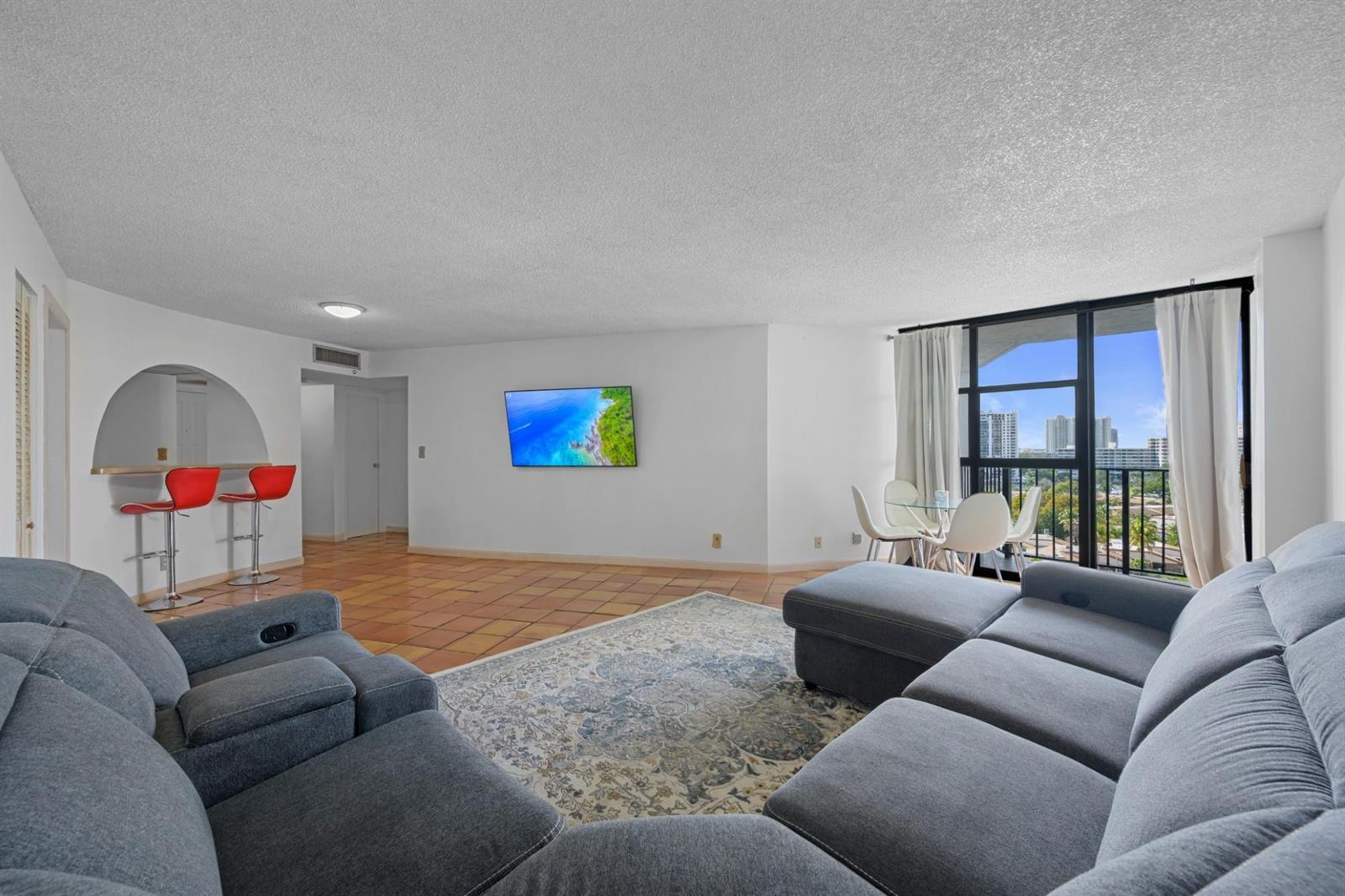A living room with a grey sectional sofa, tile floors, a wall-mounted television, and balcony access — 600 Parkview Drive, Hallandale Beach FL 33009, HALLANDALE BEACH, FL