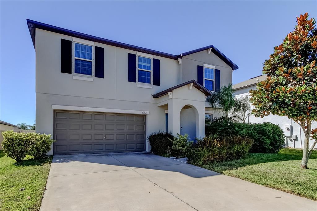 A two-story suburban house with a beige stucco exterior, blue shutters, and a concrete driveway — 6362 SPIDER LILY WAY, NEW PORT RICHEY, FL