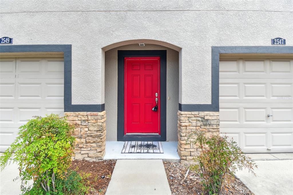 A bright red front door is centered between two beige garage doors with stone accents — 2547 LEXINGTON OAK DRIVE, BRANDON, FL