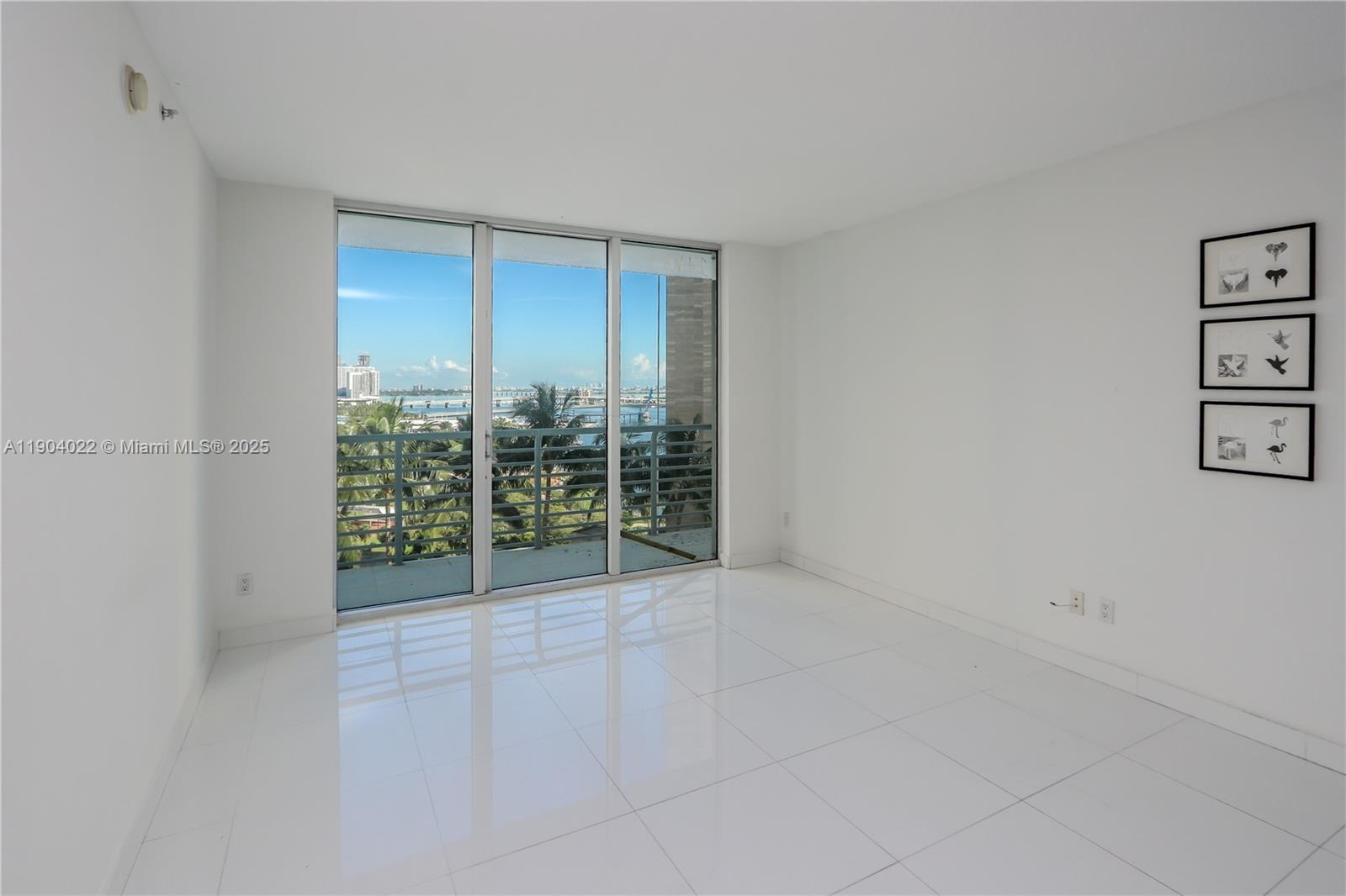 A bright, empty room features white tile floors, large sliding glass doors, and wall art — 325 S Biscayne Blvd # 1722, Miami FL 33131, MIAMI, FL