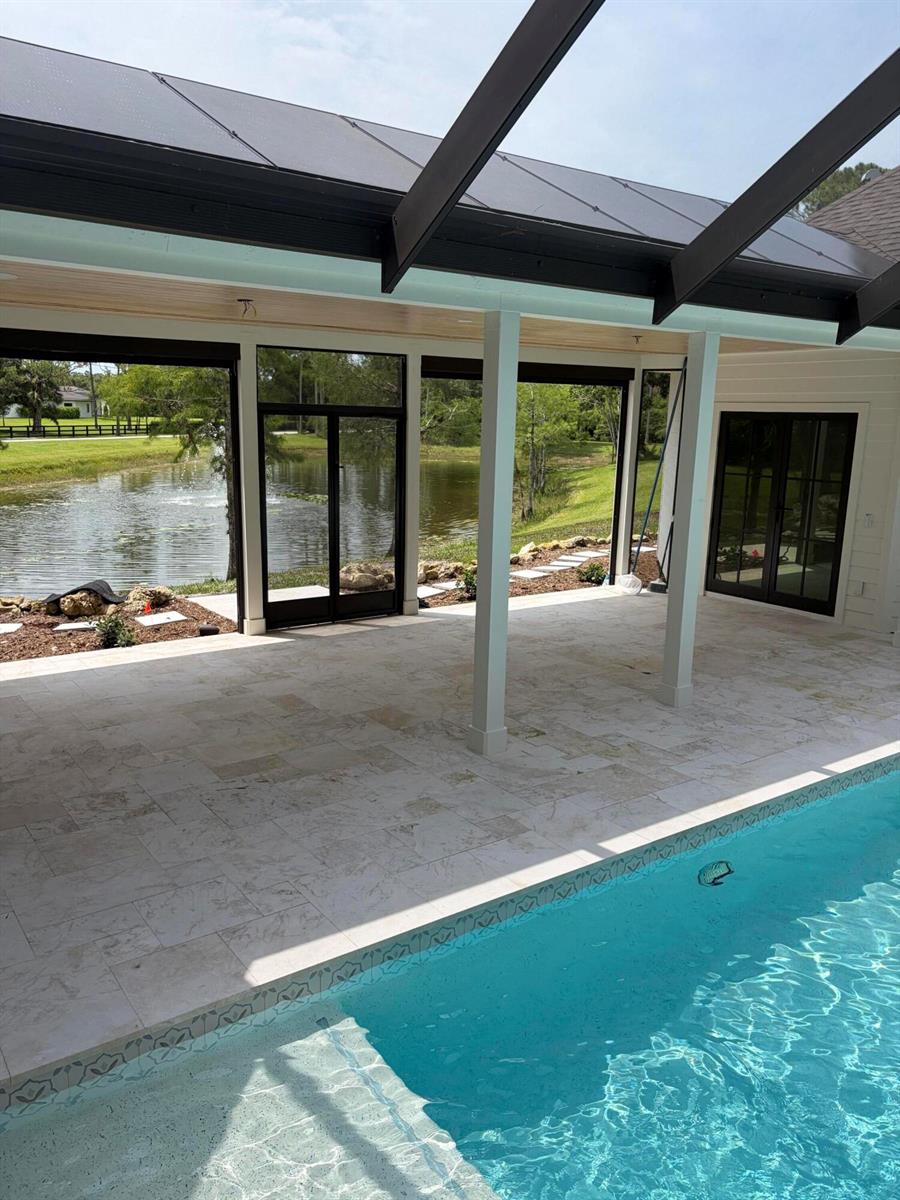 A covered patio with light stone flooring overlooks a swimming pool and a pond — 18363 Island Oak Avenue, Jupiter FL 33478, JUPITER, FL