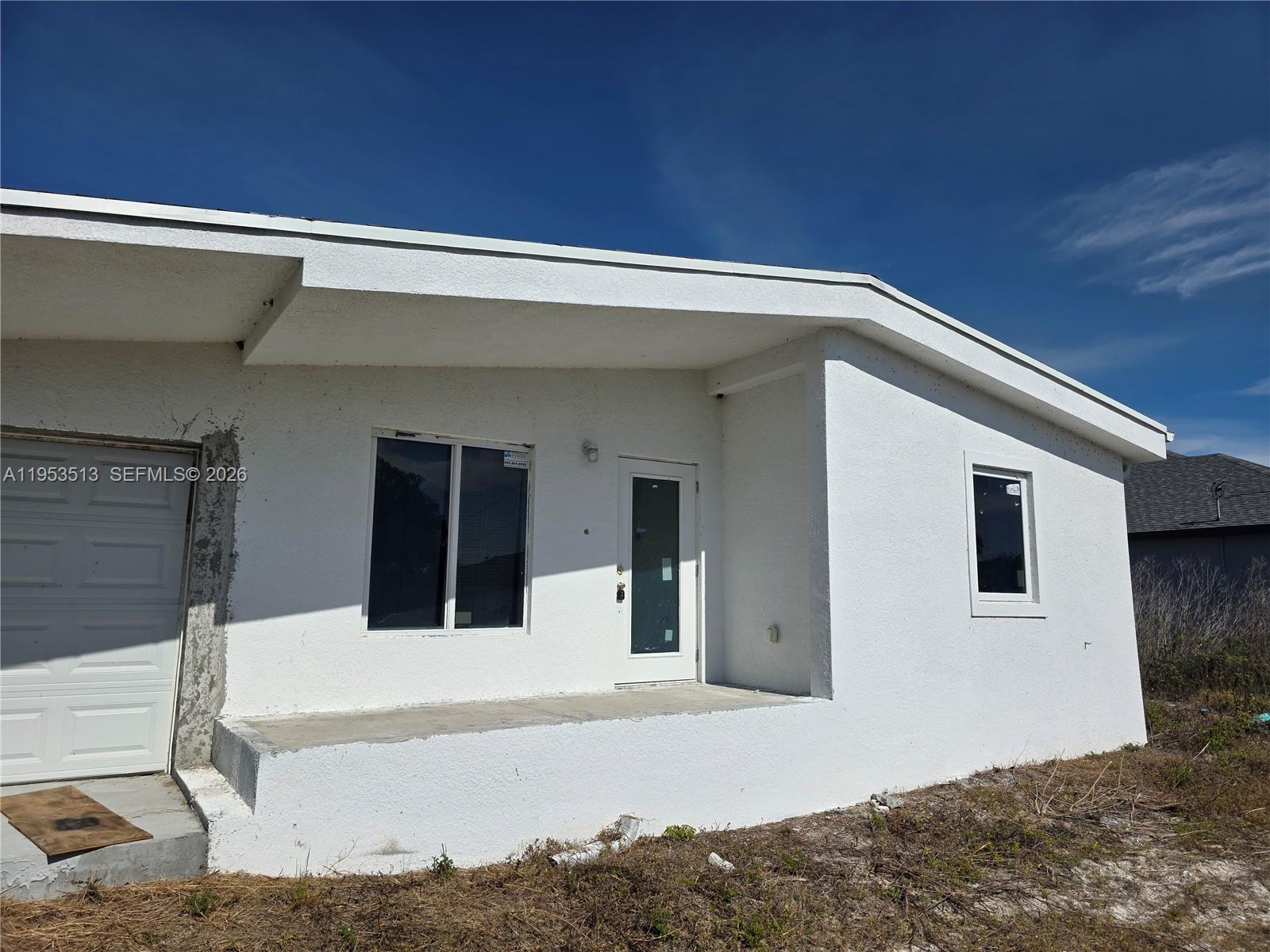 The exterior of a white, single-story house features a concrete porch and a garage door — 3030 SW 39TH ST, Lehigh Acres FL 33976, LEHIGH ACRES, FL