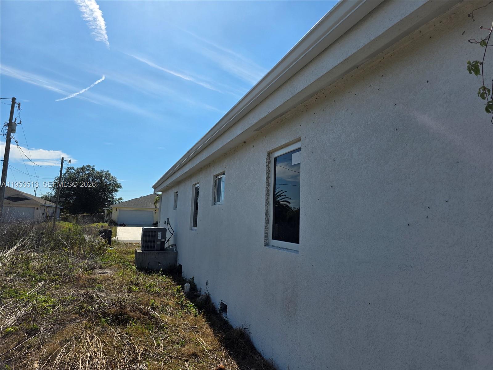 The exterior side of a white stucco house features several windows and an air conditioning unit — 3030 SW 39TH ST, Lehigh Acres FL 33976, LEHIGH ACRES, FL