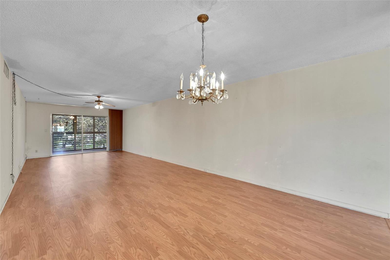 An empty living room features wood-look laminate flooring, a crystal chandelier, and a sliding glass door — 2006 Granada Drive # A3, Coconut Creek FL 33066, COCONUT CREEK, FL